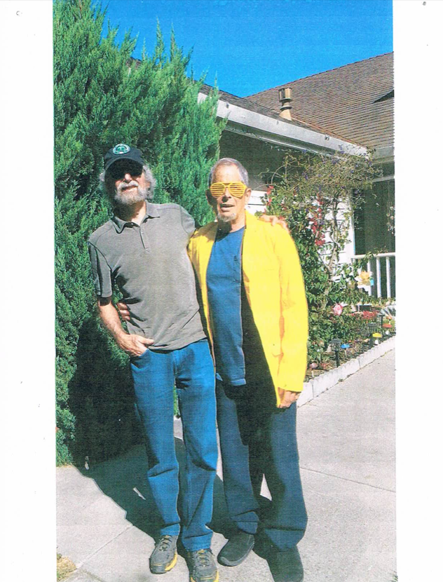 The author, Tom Wayman, with his friend, Dennis Saleh. Tom is wearing a grey t-shirt and jeans, and Dennis is wearing the bright yellow glasses and jacket.